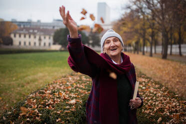 Eine glückliche ältere Frau auf einem Herbstspaziergang im Stadtpark, die Blätter wirft und in die Kamera schaut. - HPIF09108