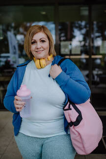 A happy overweight woman in sports clothes looking at camera outdoors in  front of fitness center