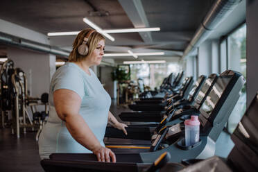 Mittlere erwachsene übergewichtige Frau mit Kopfhörern beim Laufen auf dem Laufband im Fitnessstudio - HPIF09156