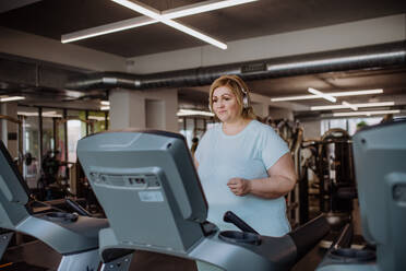 Mittlere erwachsene übergewichtige Frau mit Kopfhörern beim Laufen auf dem Laufband im Fitnessstudio - HPIF09157