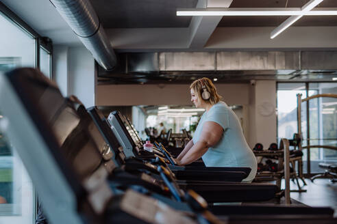 Mittlere erwachsene übergewichtige Frau mit Kopfhörern beim Laufen auf dem Laufband im Fitnessstudio - HPIF09158