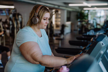 Mittlere erwachsene übergewichtige Frau mit Kopfhörern beim Laufen auf dem Laufband im Fitnessstudio - HPIF09159
