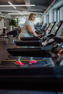 Mid adult overweight woman with headphones running on treadmill in gym - HPIF09160