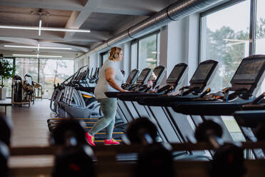 Mid adult overweight woman with headphones running on treadmill in gym - HPIF09162