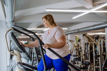 Eine glückliche übergewichtige Frau im mittleren Erwachsenenalter, die in einem Fitnessstudio auf einem Stepper trainiert - HPIF09163