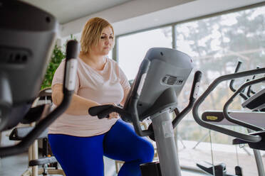 Eine glückliche übergewichtige Frau im mittleren Erwachsenenalter, die in einem Fitnessstudio auf einem Stepper trainiert - HPIF09164