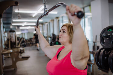 A mid adult plus size woman working out on a lat pulldown machine indoors in gym - HPIF09170