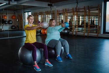 Eine übergewichtige Frau mit Hanteln auf fintess Ball sitzen und die Ausübung mit Personal Trainer im Fitnessstudio - HPIF09195