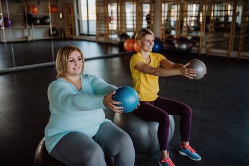 Eine glückliche übergewichtige Frau, die auf einem Fintess-Ball sitzt und mit einem Personal Trainer im Fitnessstudio trainiert - HPIF09199