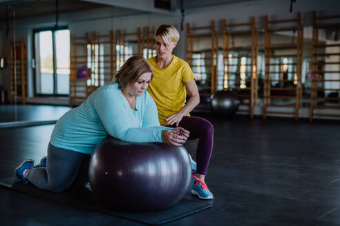 Eine glückliche übergewichtige Frau, die mit einem Personal Trainer auf einem Fintess-Ball im Fitnessstudio trainiert - HPIF09200