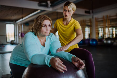 Eine glückliche übergewichtige Frau, die mit einem Personal Trainer auf einem Fintess-Ball im Fitnessstudio trainiert - HPIF09201