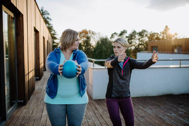 Eine übergewichtige Frau hält einen Ball und trainiert mit einem Personal Trainer im Freien auf der Terrasse eines Fitnessstudios. - HPIF09203