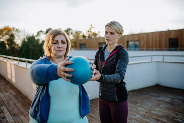 Eine übergewichtige Frau hält einen Ball und trainiert mit einem Personal Trainer im Freien auf der Terrasse eines Fitnessstudios. - HPIF09204