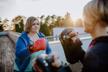 An overweight woman training boxing with personal trainer outdoors on terrace. - HPIF09208