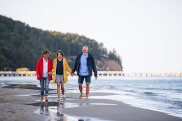 A senior couple holding hands with their preteen granddaughter and walking on sandy beach. - HPIF09220