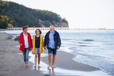 A senior couple holding hands with their preteen granddaughter and walking on sandy beach. - HPIF09221