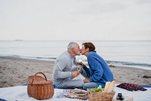 Ein glückliches, verliebtes älteres Paar sitzt auf einer Decke und küsst sich beim Picknick im Freien am Strand. - HPIF09261