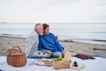 A happy senior couple in love sitting on blanket and having picnic outdoors on beach by sea. - HPIF09263