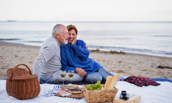 Ein glückliches, verliebtes älteres Paar sitzt auf einer Decke und macht ein Picknick im Freien am Strand am Meer. - HPIF09264
