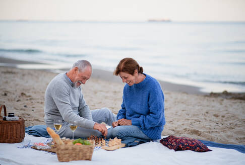 Ein glückliches, verliebtes älteres Paar, das auf einer Decke sitzt, Wein trinkt und draußen am Strand Schach spielt. - HPIF09268