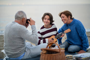Ein glückliches älteres Paar mit einer Enkelin, die auf einer Decke sitzt und ein Picknick im Freien am Strand am Meer macht. - HPIF09269
