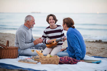 Ein glückliches älteres Paar mit einer Enkelin, die auf einer Decke sitzt und ein Picknick im Freien am Strand am Meer macht. - HPIF09270