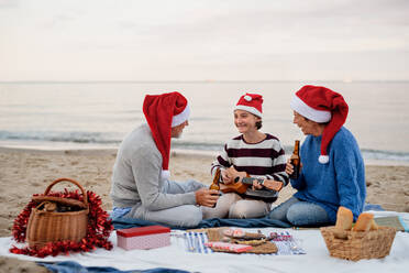 Ein glückliches älteres Paar mit einer Enkelin, die auf einer Decke sitzt und ein Picknick im Freien am Strand am Meer macht. - HPIF09272