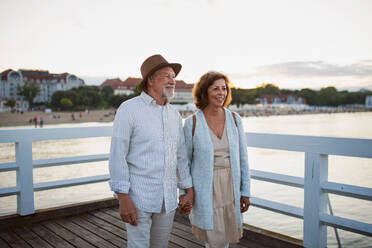 Ein glückliches älteres Paar, das sich bei einem Spaziergang im Freien auf dem Pier am Meer die Hände hält und die Aussicht genießt. - HPIF09276