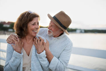 A happy senior couple hugging outdoors on pier by sea, looking at each other. - HPIF09278