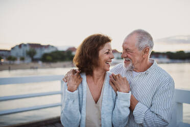 A happy senior couple hugging outdoors on pier by sea, looking at each other. - HPIF09280