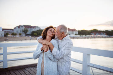 A happy senior couple hugging outdoors on pier by sea, looking at each other. - HPIF09282