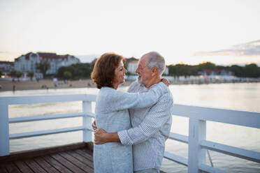 A happy senior couple hugging outdoors on pier by sea, looking at each other. - HPIF09283