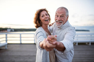 Ein glückliches älteres Paar tanzt im Freien auf einem Pier am Meer und schaut in die Kamera. - HPIF09304