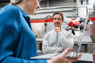 Smiling engineer with hand on chin looking at colleague in robot factory - DIGF19836