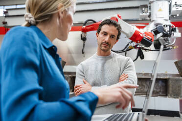 Colleague explaining engineer with arms crossed in robot factory - DIGF19837