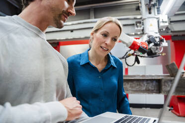 Mature engineer looking at laptop held by colleague in robot factory - DIGF19838