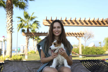 Glückliche junge Frau mit Hund auf Hängematte im Park sitzend - SYEF00239