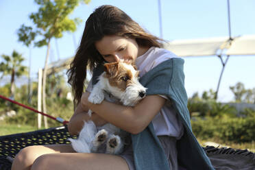 Glückliche junge Frau mit Jack Russell Terrier auf Hängematte sitzend an einem sonnigen Tag - SYEF00244