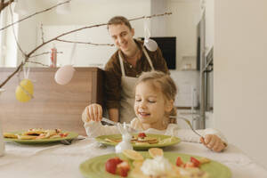 Lächelndes Mädchen isst Pfannkuchen auf einem Tisch zu Hause - VIVF00432