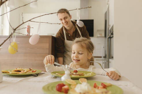 Smiling girl eating pancakes on table at home - VIVF00432