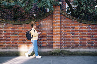 Young woman using smart phone and walking on footpath by brick wall - AMWF01189