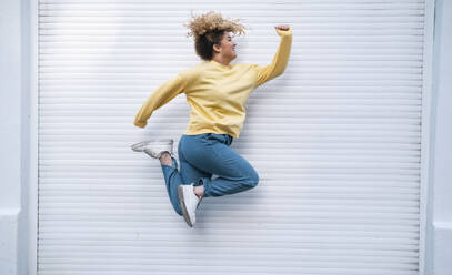 Carefree young woman jumping by closed white shutter - AMWF01190