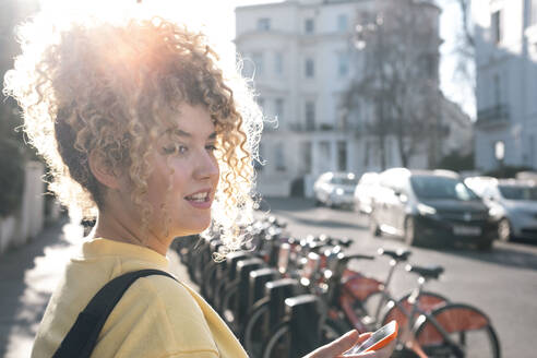 Junge Frau mit Smartphone an einer Fahrradparkstation an einem sonnigen Tag - AMWF01199