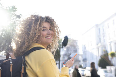 Smiling woman with backpack and smart phone on sunny day - AMWF01200