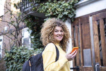 Smiling young woman with curly hair holding smart phone outside house - AMWF01204