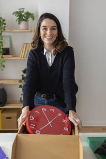 Smiling businesswoman with wall clock in box standing at desk - XLGF03261