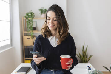 Smiling businesswoman with coffee cup using smart phone in office - XLGF03282