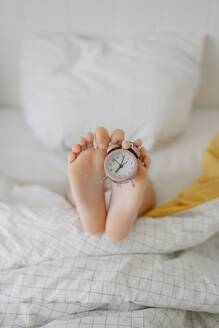 Feet of girl with alarm clock on bed at home - SSYF00115