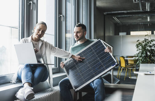 Geschäftsfrau erklärt Kollegen, die ein Solarpanel im Büro halten - UUF28414