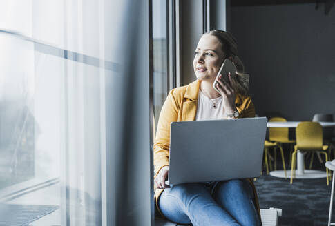 Lächelnde Geschäftsfrau, die am Fenster sitzend mit einem Laptop im Büro über ein Smartphone spricht - UUF28454